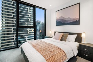 A modern bedroom with a large bed, a city view, and a mountain landscape painting. - Stylish City 2 Bedroom Gem (Melbourne)
