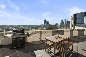 Eine Dachterrasse mit Grill, Tisch und Stühlen mit Blick auf eine Stadtlandschaft.