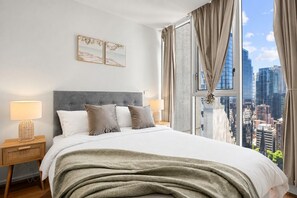 Une chambre moderne avec un grand lit, deux lampes de chevet, une vue sur la ville et une tête de lit grise.