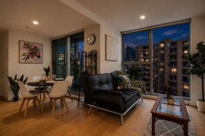A modern living room with a dining area, a black leather sofa, a dining table with chairs, and a large window offering a city view. - Urban Jungle 2 Bedroom Escape (Melbourne)