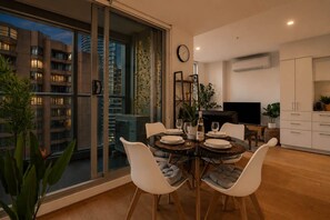 A modern dining area with a round table, white chairs, and a city view through large windows. - Urban Jungle 2 Bedroom Escape (Melbourne)