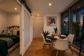 A modern bedroom with a bed, a dining area with a table and chairs, and a city view through large windows. - Urban Jungle 2 Bedroom Escape (Melbourne)