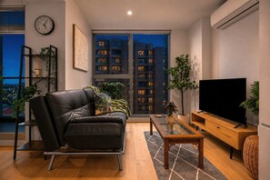 A modern living room with a leather sofa, a flat-screen TV, and a large window overlooking a cityscape at night. - Urban Jungle 2 Bedroom Escape (Melbourne)