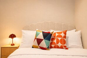 Ein Bett mit weißer Bettwäsche, zwei Kissen mit geometrischen Mustern, ein Nachttisch aus Holz mit einer roten Lampe und ein beige gepolstertes Kopfteil.