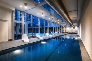 A modern indoor swimming pool with clear glass walls, lounge chairs, and ceiling lights. - Retro City 3 Bedroom Retreat (Melbourne)