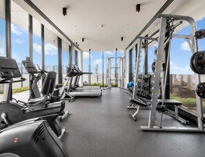 A gym with cardio machines, weightlifting equipment, and large windows offering a view of the sky. - Modern City 2 Bedroom (Melbourne)