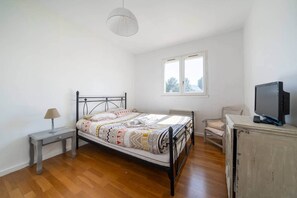 Un dormitorio con cama, mesita de noche, lámpara, silla, televisor y ventana.