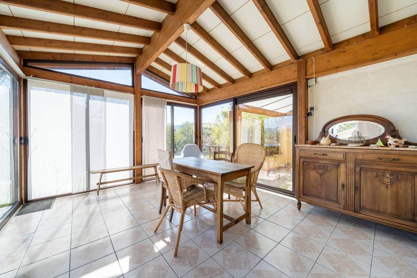 Une pièce dotée d’un plafond en bois, d’une table à manger avec des chaises, d’un meuble contenant des objets décoratifs et de grandes fenêtres offrant une vue sur l’extérieur.