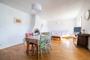 Un área de comedor con una mesa y sillas, un sofá, una chimenea y un televisor en una habitación luminosa con piso de madera.