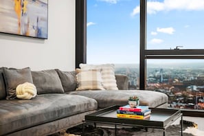 A modern living room with a grey sofa, a coffee table with books, and a large window offering a city view. - Level 61 City View 3 Bedroom (Melbourne)