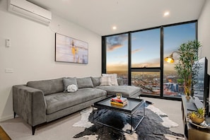 A modern living room with a grey sofa, a glass coffee table, and a city view through large windows. - Level 61 City View 3 Bedroom (Melbourne)