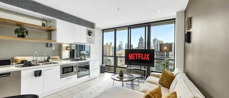 A modern living room with a sofa, a TV displaying 'NETFLIX,' and a city view through large windows.