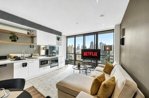 A modern living room with a sofa, a TV displaying 'NETFLIX,' and a city view through large windows. - River View Southbank Apartment (Melbourne)