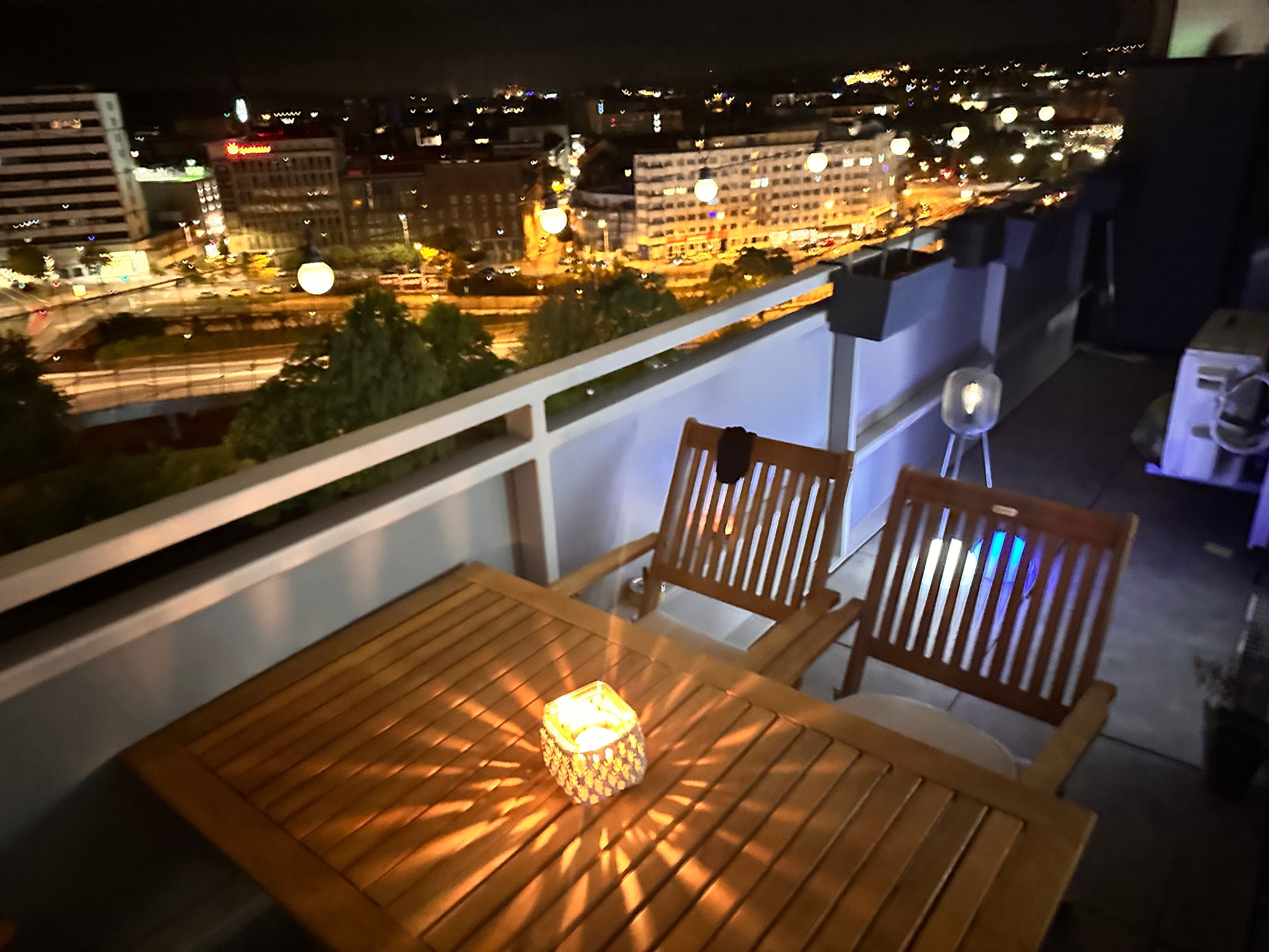 Ein Balkon mit Holzmöbeln und einem Tisch mit Blick auf eine beleuchtete Stadtlandschaft bei Nacht.