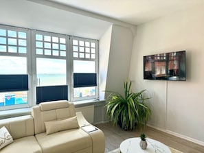 Ein modernes Wohnzimmer mit einem beigefarbenen Sofa, einer großen Topfpflanze, einem an der Wand befestigten Flachbildfernseher und Meerblick durch große Fenster.