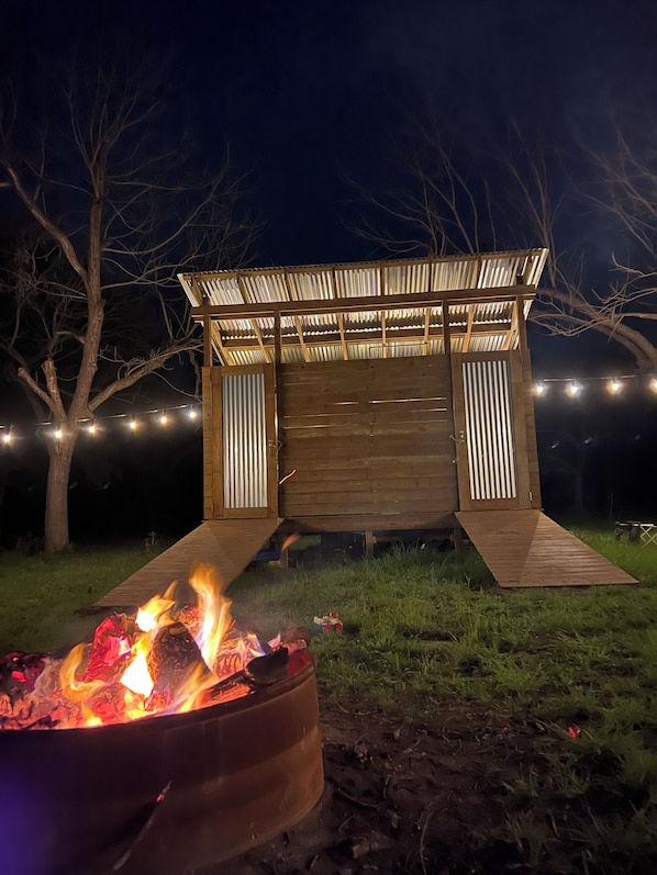 A wooden structure with a ramp leading to a covered entrance, surrounded by trees and a campfire.