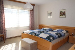 A bedroom with a wooden bed, a bedside table, a nightstand, a bench, a radiator, a window with curtains, and two framed pictures on the wall. - Vacation Rental on the Ilsenbach, 105 m², Right in the Heart of the Village (Lügde)