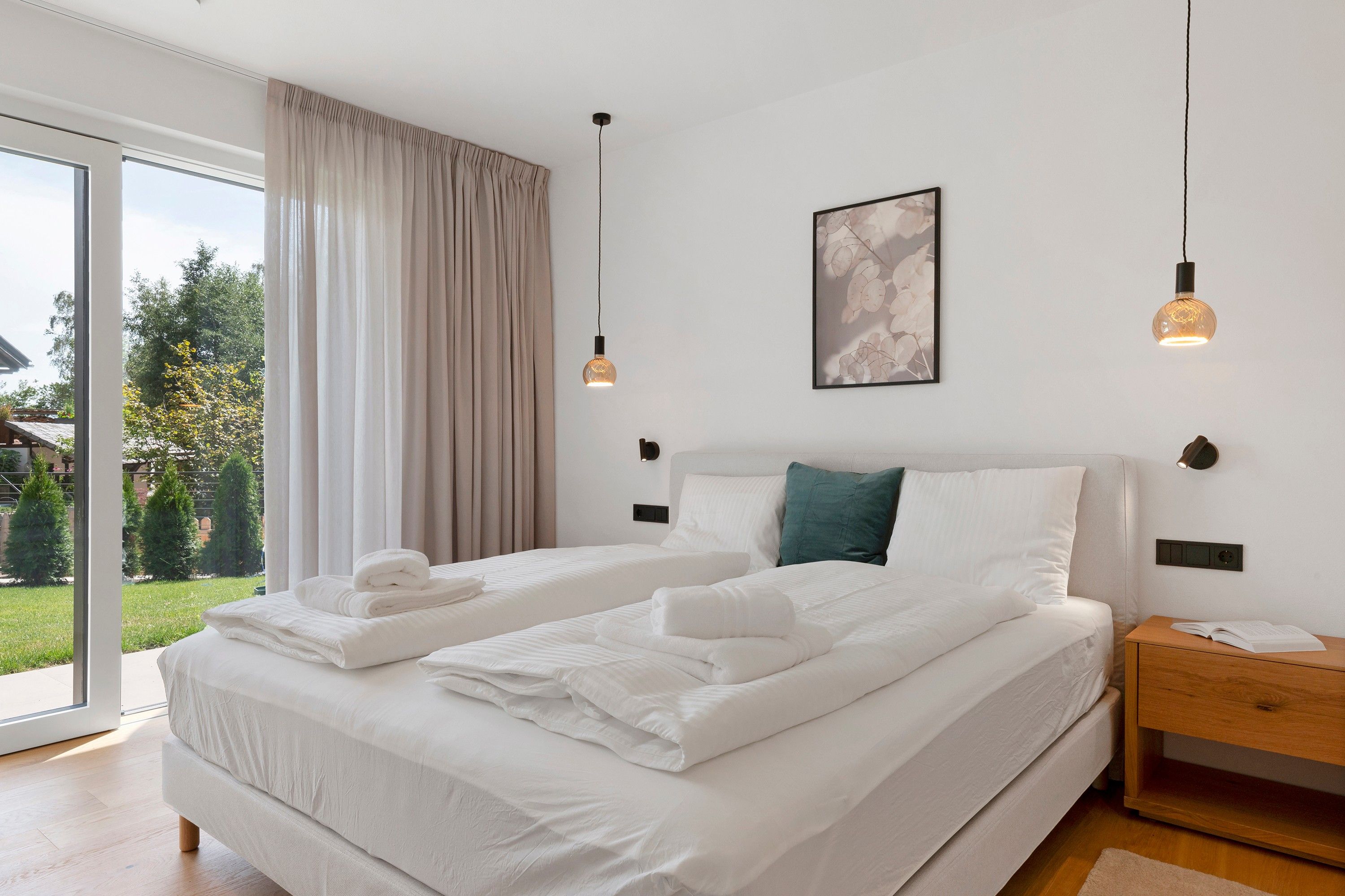 Ein modernes Schlafzimmer mit einem großen Bett, zwei Nachttischen, einem kleinen Tischchen, einem gerahmten Bild und Blick ins Freie.