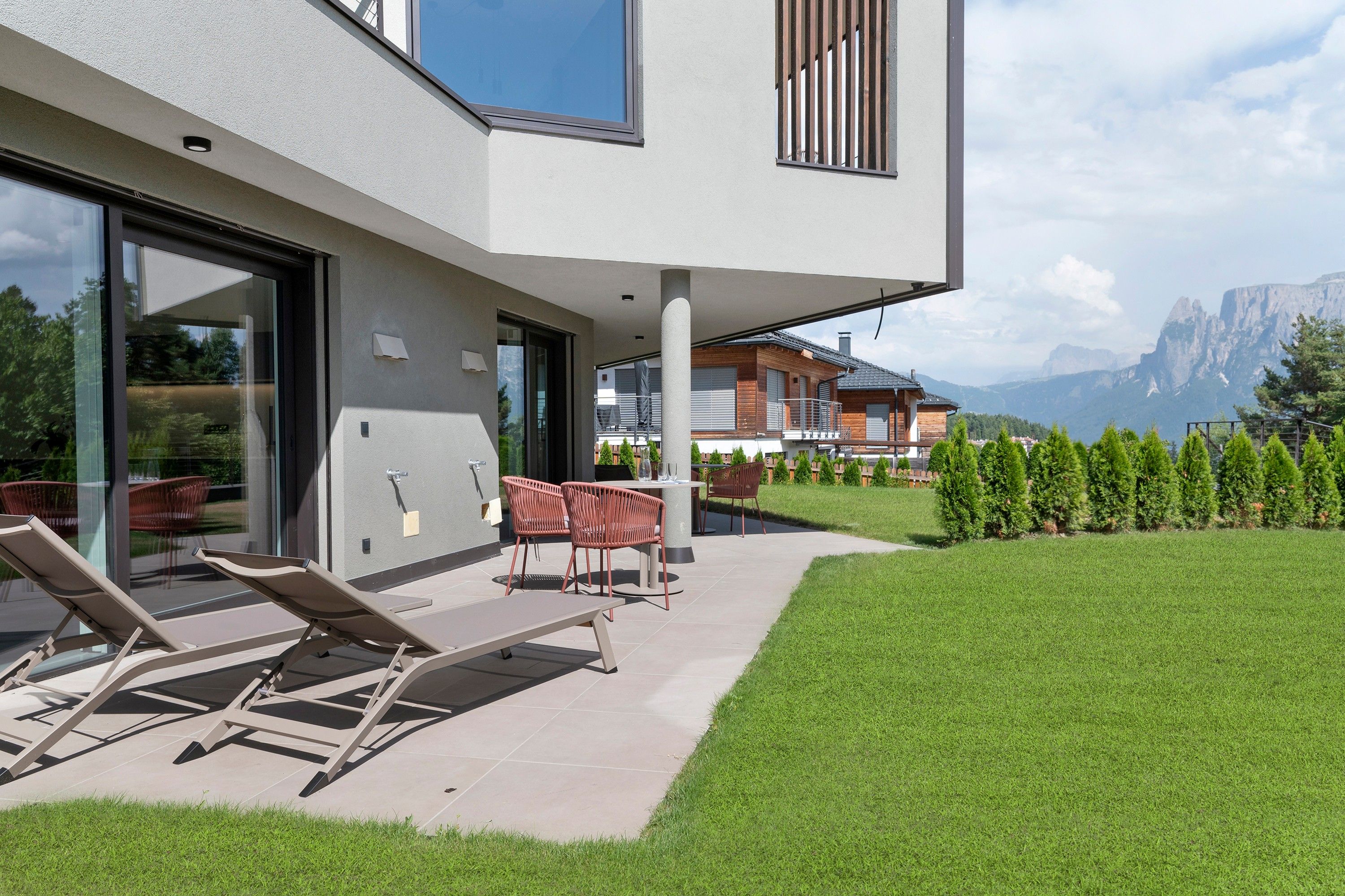 A modern outdoor seating area with lounge chairs and a table, a glass door entrance, and a view of a mountain landscape.