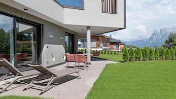 A modern outdoor seating area with lounge chairs and a table, a glass door entrance, and a view of a mountain landscape.