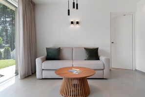 Ein modernes Wohnzimmer mit einer Couch, einem runden Holztisch und Blick auf einen Wald durch ein großes Fenster.