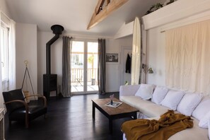 Ein Wohnzimmer mit einer Couch, einem hölzernen Couchtisch, einem schwarzen Ledersessel, einem schwarzen Kaminofen und Blick ins Freie durch eine Glastür.