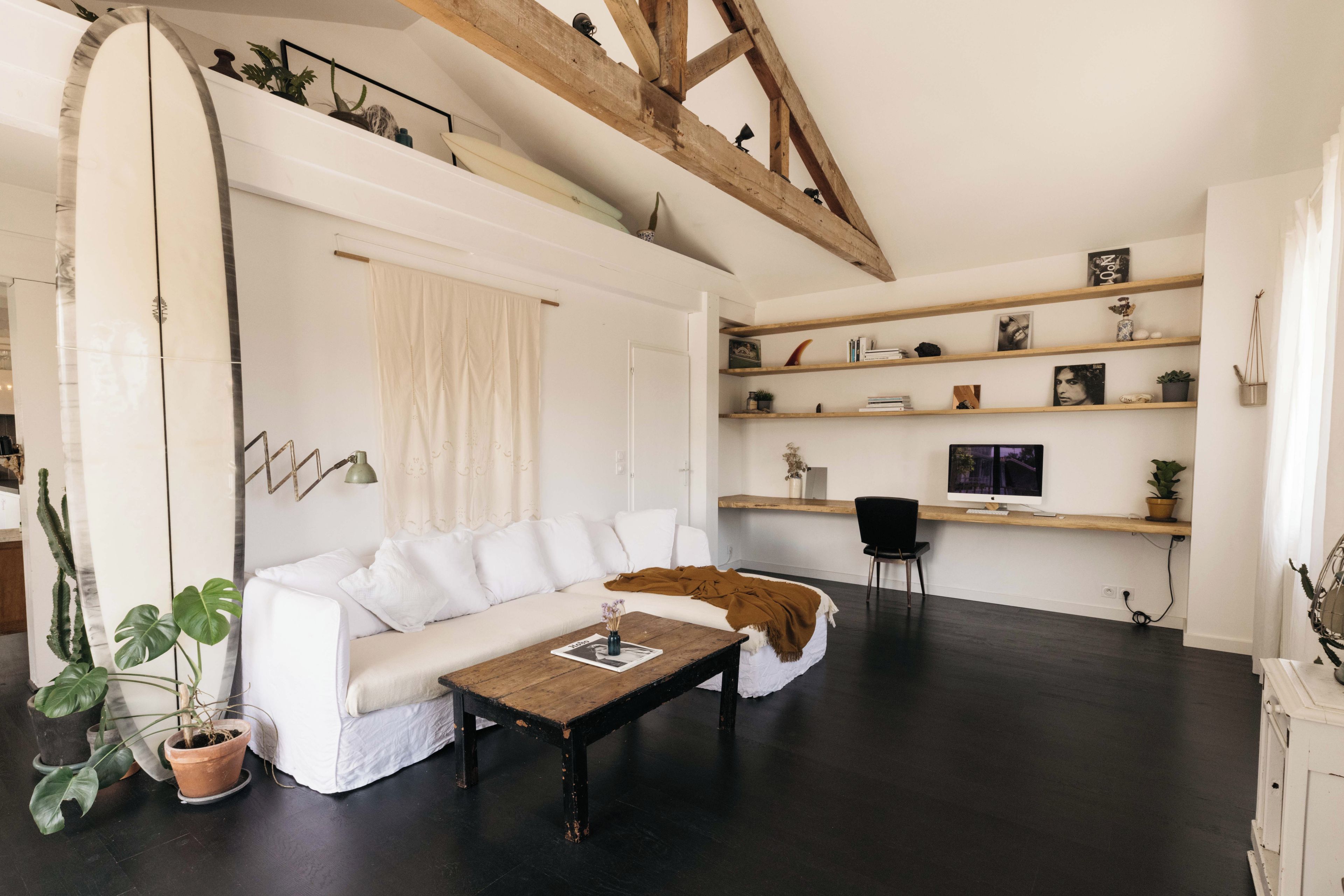 A modern living room with a white sofa, a coffee table, and a surfboard leaning against the wall. There are shelves with various decorative items and a computer monitor on a desk.