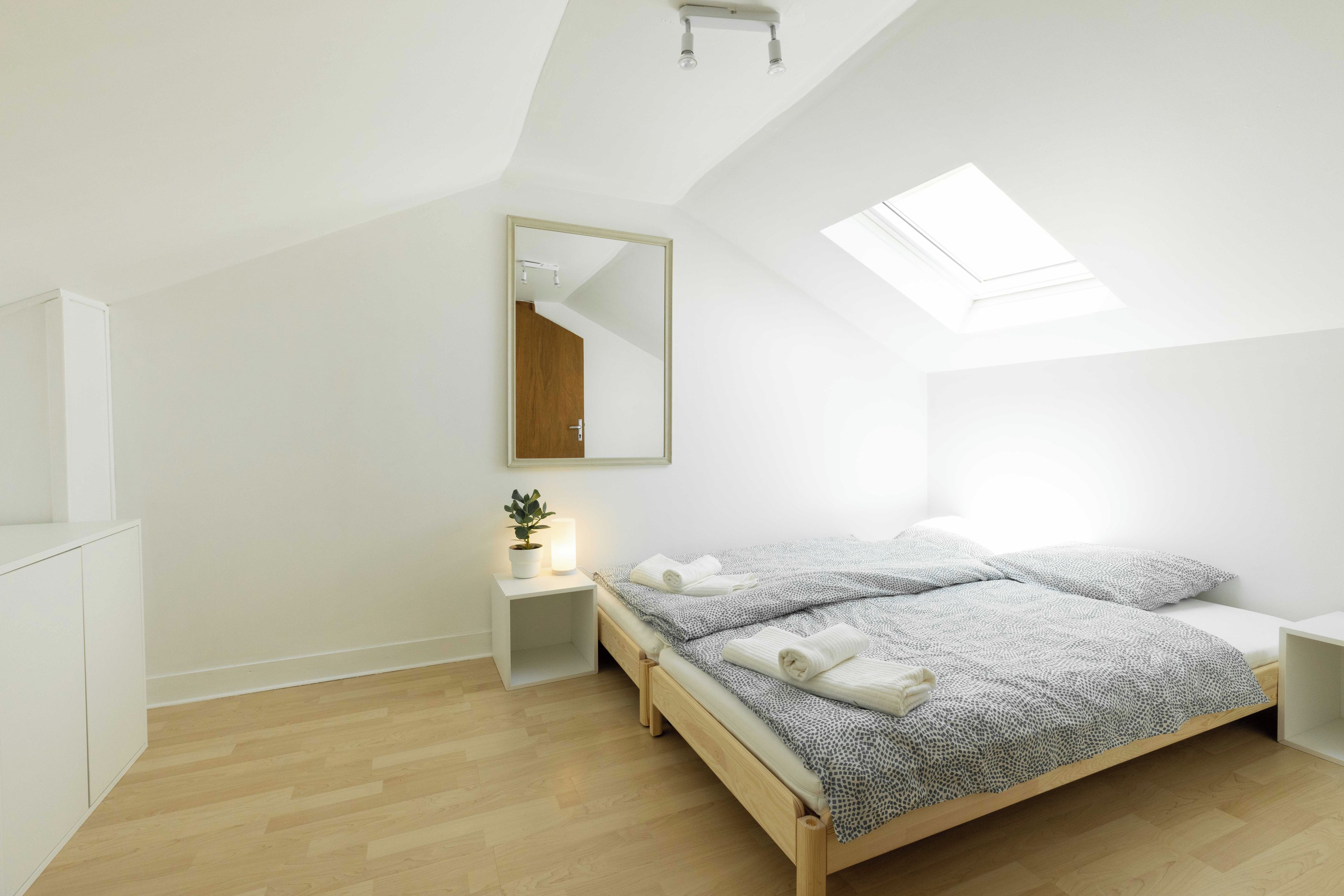 A modern bedroom with a sloped ceiling, a skylight, a bedside table with a lamp, and a bed with a patterned bedspread and rolled towels.