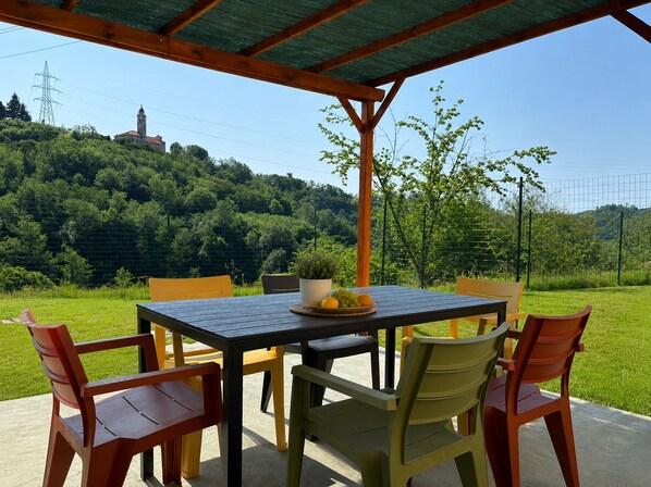 An outdoor dining area with a table and chairs, a potted plant, and a view of a hill with a tower.
