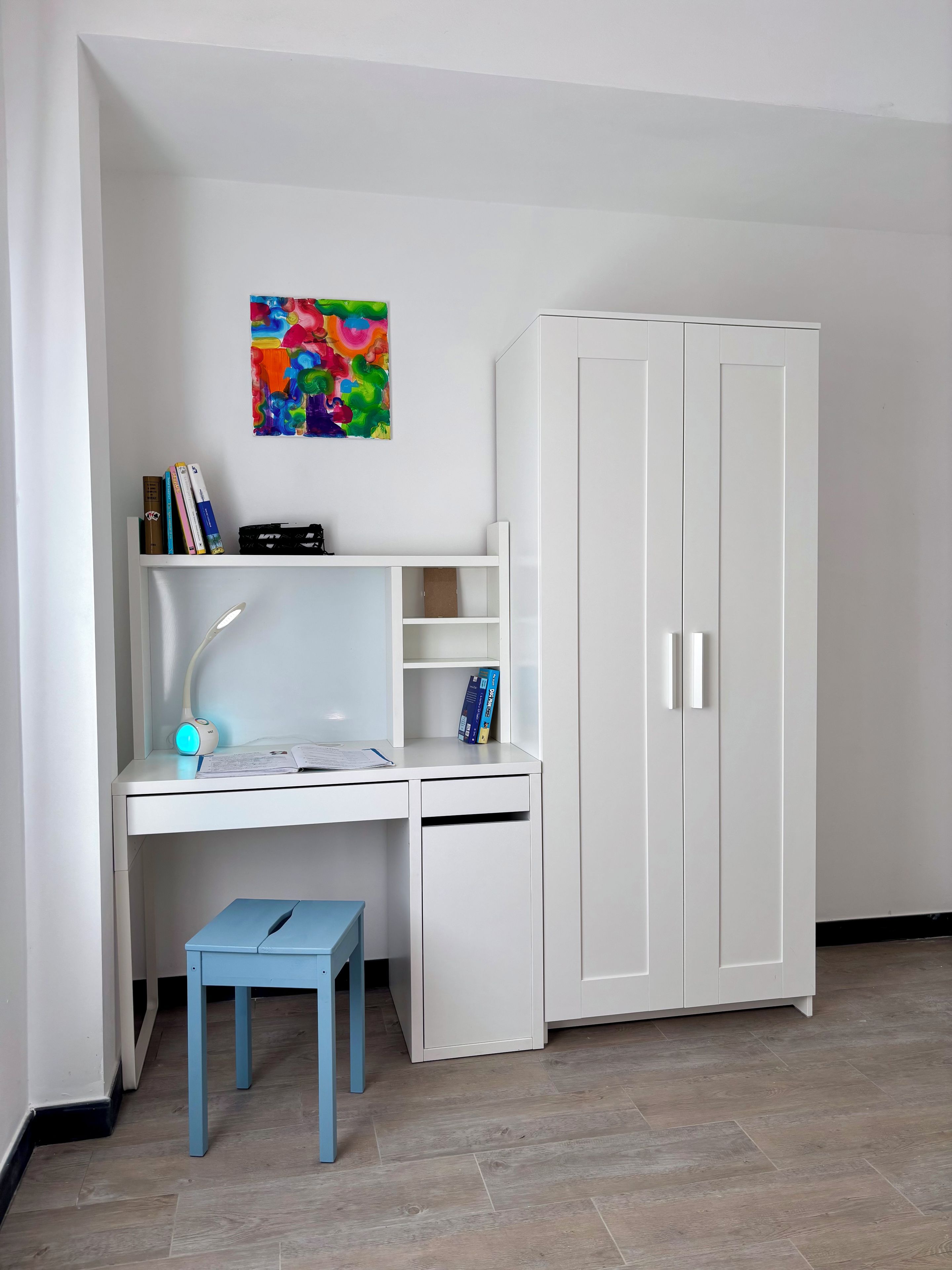 Ein Zimmer mit einem weißen Schreibtisch, einem blauen Hocker, einem weißen Kleiderschrank und einem farbenfrohen abstrakten Gemälde an der Wand.