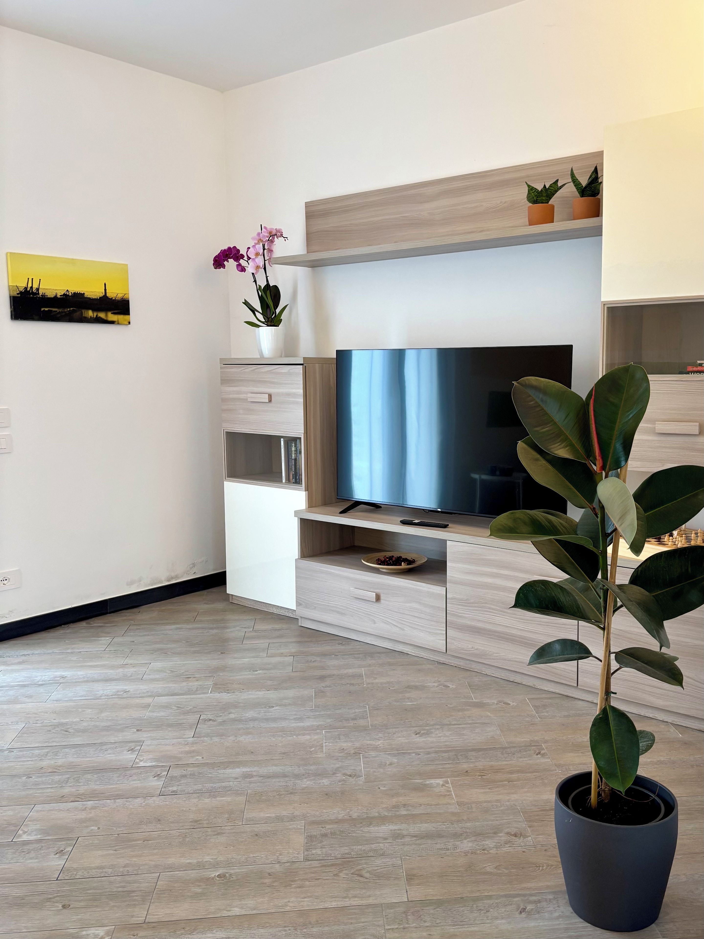 A modern living room with a flat-screen TV, wooden flooring, a potted plant, and a painting on the wall.