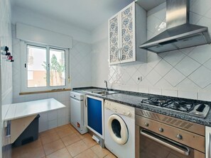 A compact kitchen with a built-in washer, a gas stove, and a window with a view of palm trees. - Apartment 'Quinta Da Gomeira 68' with shared pool, Wi-Fi and air conditioning (Tavira)