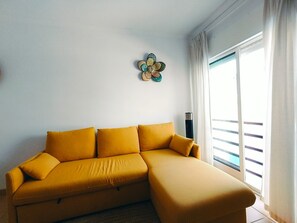 A yellow sofa with cushions in a room with a large window and a decorative wall piece. - Apartment 'Quinta Da Gomeira 68' with shared pool, Wi-Fi and air conditioning (Tavira)