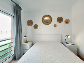 A bedroom with a large bed, two bedside tables with lamps, a window with curtains, and wall-mounted decorative items. - Apartment 'Quinta Da Gomeira 68' with shared pool, Wi-Fi and air conditioning (Tavira)