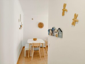 A dining area with a white table and chairs, a woven basket on the table, and wall decorations including a circular woven item and small house-shaped decorations. - Apartment 'Quinta Da Gomeira 68' with shared pool, Wi-Fi and air conditioning (Tavira)