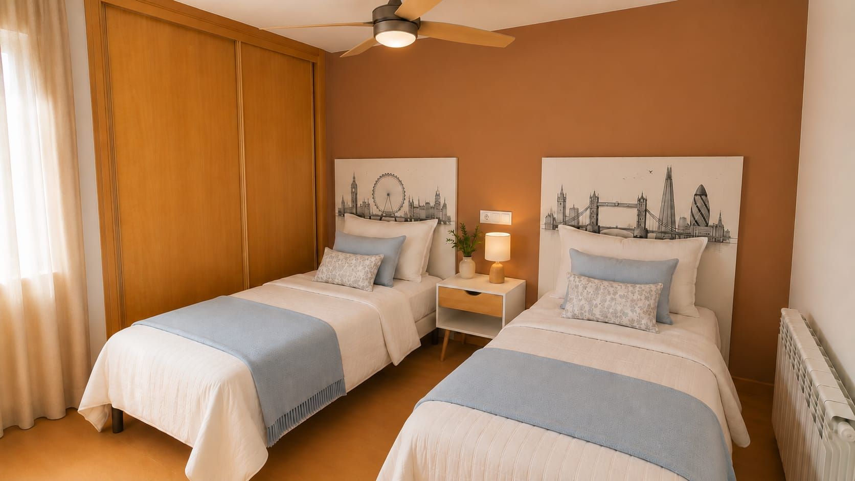 Hotellihuone, jossa on kaksi sänkyä, puinen vaatekaappi, kattotuuletin ja kaksi kaupunkimaisemaa esittävää taulua seinillä.