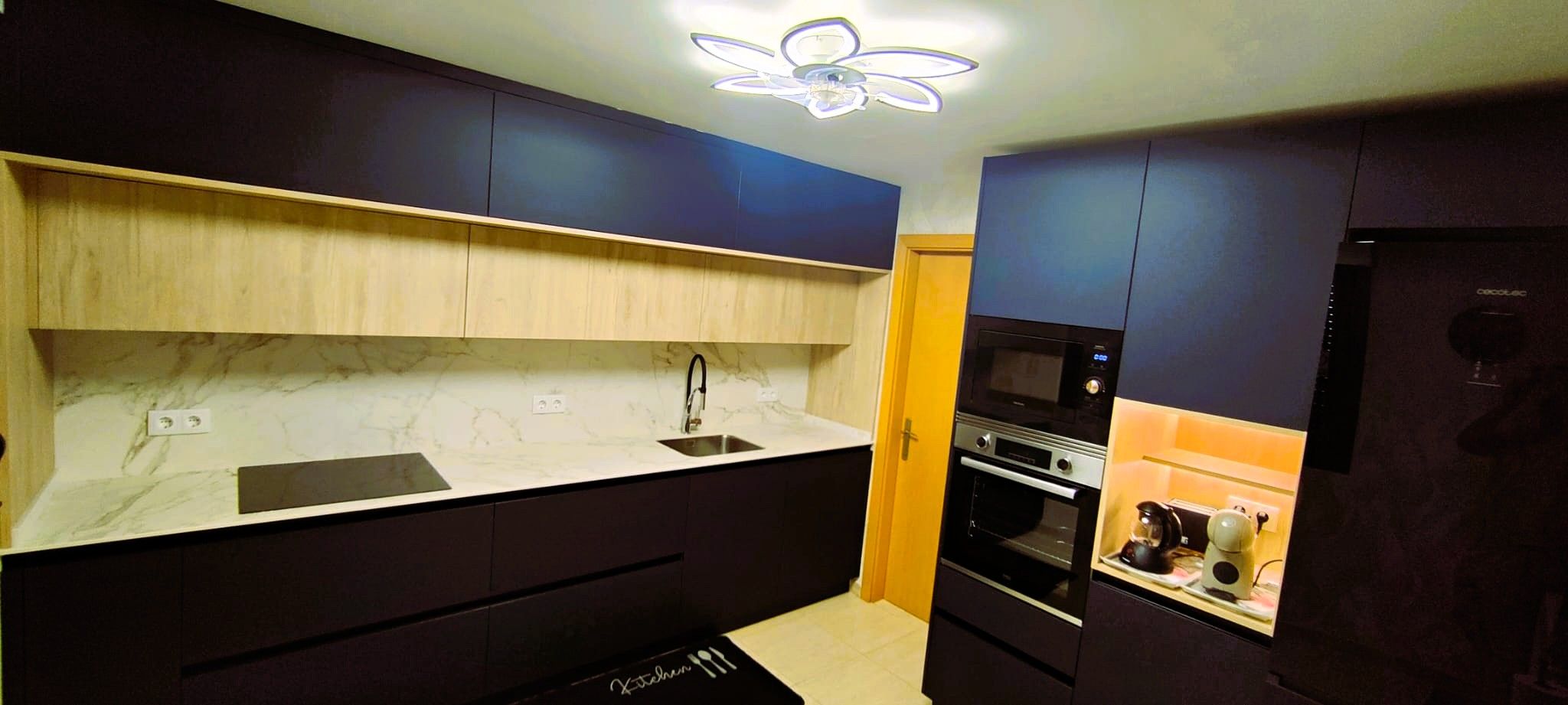 Une cuisine moderne avec des meubles de couleur sombre, un plan de travail en marbre, un four encastré et un ventilateur de plafond.