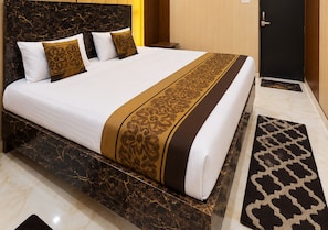 A hotel room with a large bed, a marble bedside table, a wooden door, and a patterned rug. - Hotel Utsav Kunj (Sitapur)