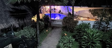 A pathway leading to a pool area with palm trees and a thatched-roof structure.
