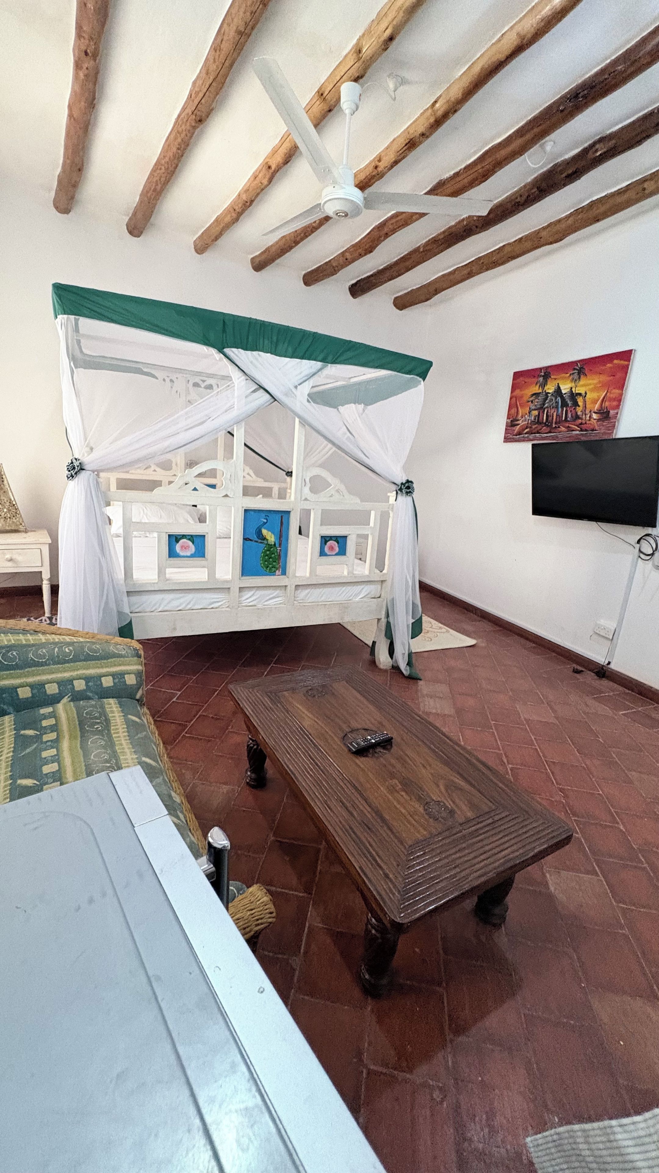 A room with a bed, a wooden table, a ceiling fan, and a painting on the wall.