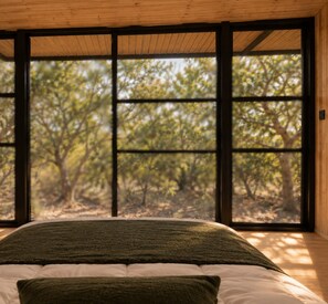 A bedroom with a large window, a bed with a green blanket, and a view of trees outside. - River and Ridge (Tierpoort)