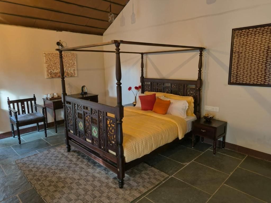 A four-poster bed with a wooden frame and a yellow bedspread, a wooden chair, a small bedside table with a lamp, and a patterned wall hanging.