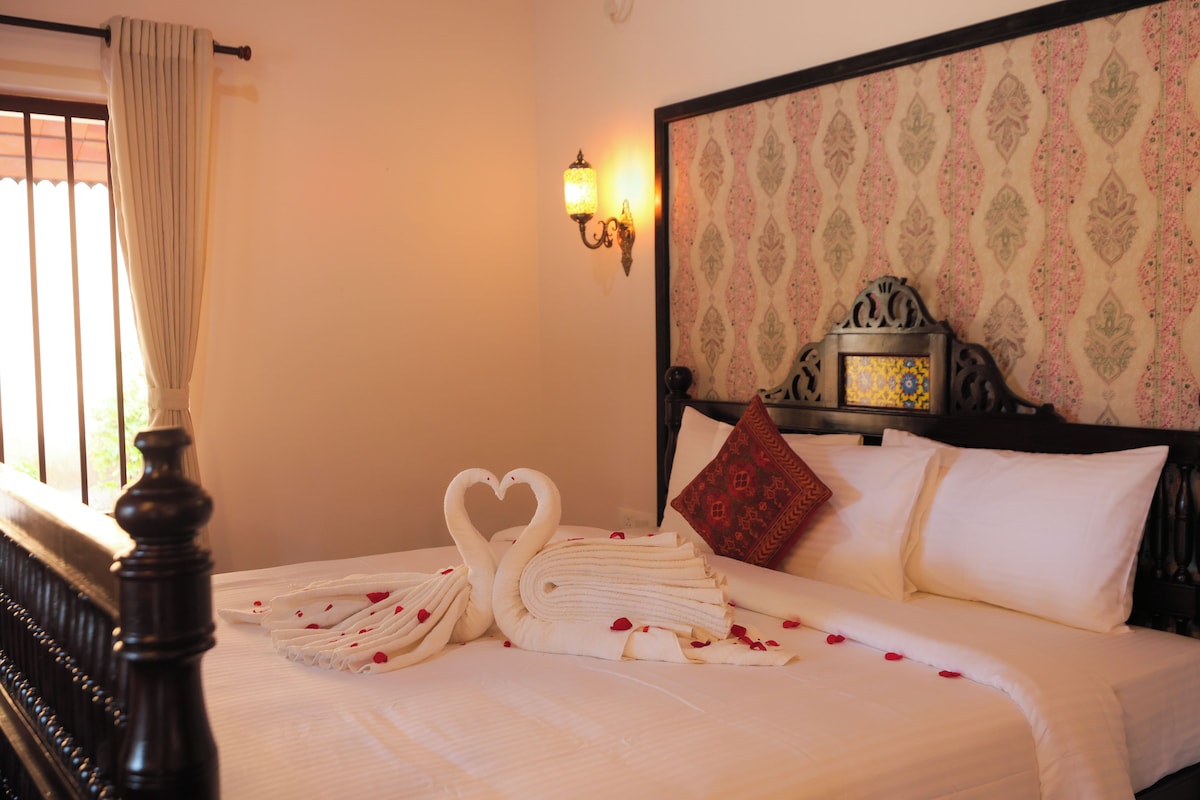Une chambre d’hôtel avec un lit, une lampe fixée au mur, une tête de lit décorative et une fenêtre avec des rideaux.