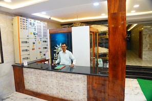 Un mostrador de recepción con un hombre detrás, un panel de control a la izquierda y una pared decorativa con un logotipo.