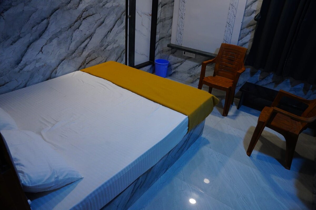 Una habitación con una cama, dos sillas de madera, un bote de basura azul y una pizarra en la pared.