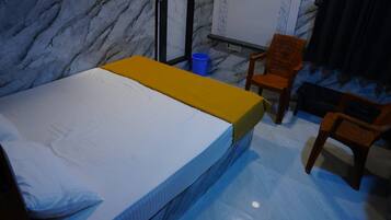 Una habitación con una cama, dos sillas de madera, un bote de basura azul y una pizarra en la pared.