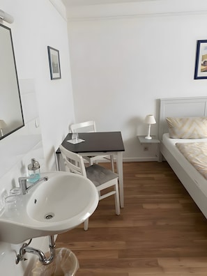Ein kleiner, sauberer Raum mit Waschbecken, Tisch, Stuhl, Bett und Nachttisch mit Lampe.