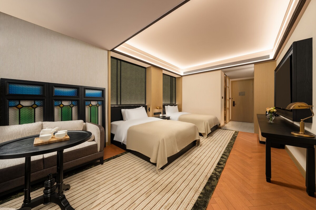 A hotel room with a sofa, two beds, a desk, and a view of the cityscape through the window.