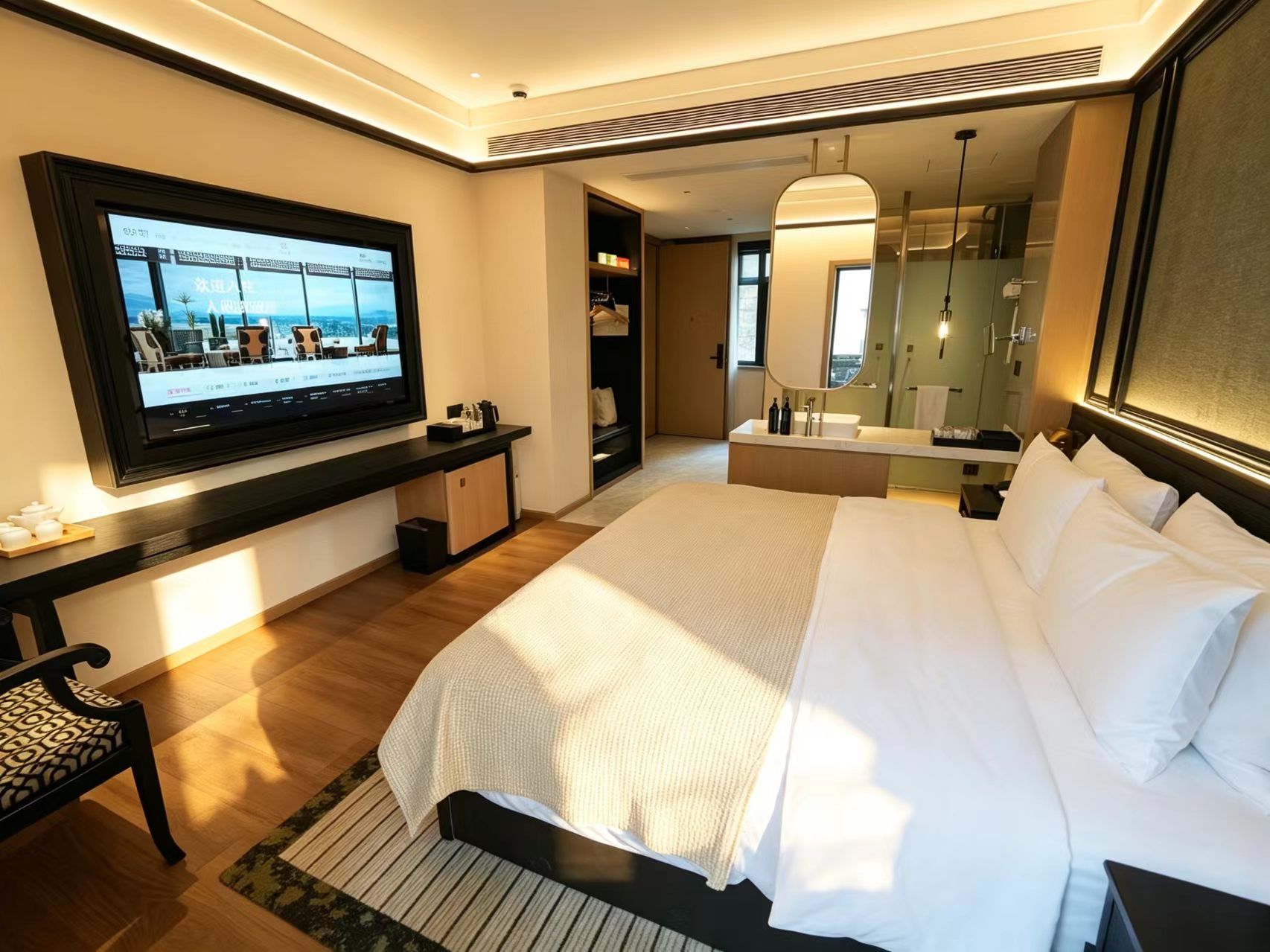 A hotel room with a bed, a TV mounted on the wall, a bedside table, a mirror, and a bathroom with a shower and sink.