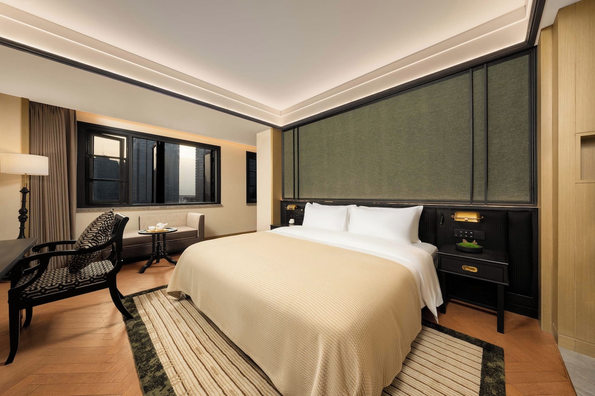 A hotel room with a large bed, a desk, a chair, a window, and a nightstand with a lamp.